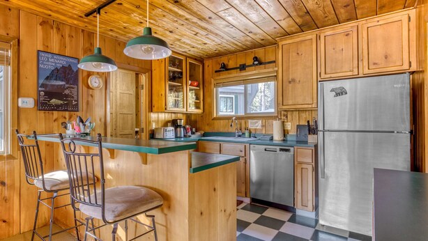 A wooden cabin kitchen with a central island, bar stools, a refrigerator, and a window overlooking the forest. - Cozy pine interior home in the heart of Government Camp (Government Camp)