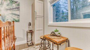 A room with a wooden crib, a small wooden table with a plant, a white cabinet, and a window with a view of trees.