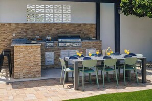 An outdoor kitchen with a brick wall, a grill, and a dining table set for four with plates and glasses. - Modern villa in Safir (Noord) with private pool and outdoor kitchen. Minutes from Eagle & Palm Beach. Perfect for relaxing, dining outside, and enjoying Aruba’s best beaches in comfort and style. (Noord)