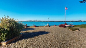 En strand med småsten, en segelbåt och en amerikansk flagga, en klarblå himmel och avlägsna kullar.