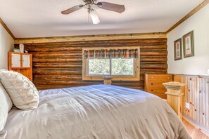 Ein Schlafzimmer in einer Blockhütte mit einem Bett, Holzwänden, einem Fenster mit Vorhängen, einem Deckenventilator und einer Holzkommode.