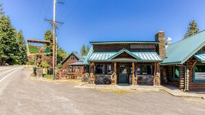 A rustic log cabin with a green roof, a stone chimney, and a wooden porch with benches. - Mountain & valley-view dog-friendly cabin with courtyard, partial AC, and WiFi (White Salmon)