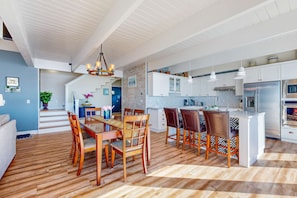A modern kitchen with white cabinets, a central island, and a dining area with wooden chairs. - Oceanfront Home with Private Hot Tub, Deck Grill & Uninterrupted Pacific Views (South Beach)