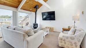 Ein Wohnzimmer mit einer Couch, einem gemusterten Sessel, einem Fernseher, einem Kamin und Blick ins Freie.