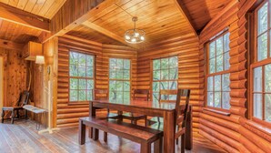 A log cabin interior with a wooden dining table, chairs, and large windows.
