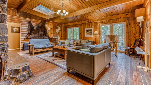 A log cabin interior with a stone fireplace, wooden furniture, and a large bear head mounted on the wall.
