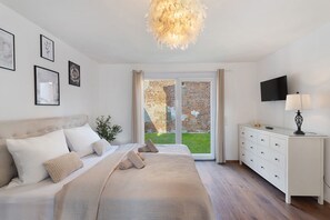 Un dormitorio con cama, cómoda, televisión y vista a un muro de ladrillo y follaje exterior.