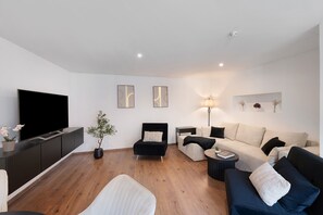 Ein modernes Wohnzimmer mit Flachbildfernseher, einem schwarzen Sessel, einem beigefarbenen Sofa, einem runden Couchtisch und Holzboden.