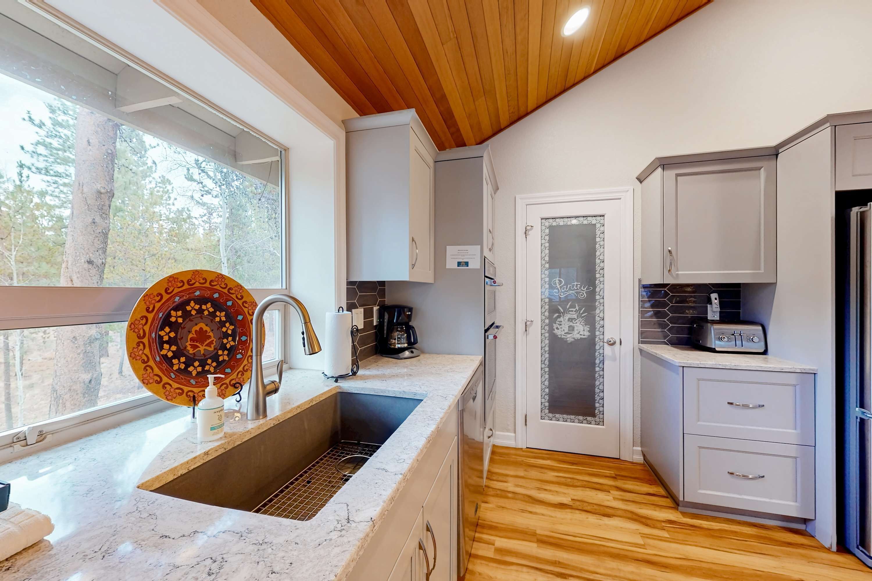 A kitchen with a sink, countertop, cabinets, and a window with a view of trees.