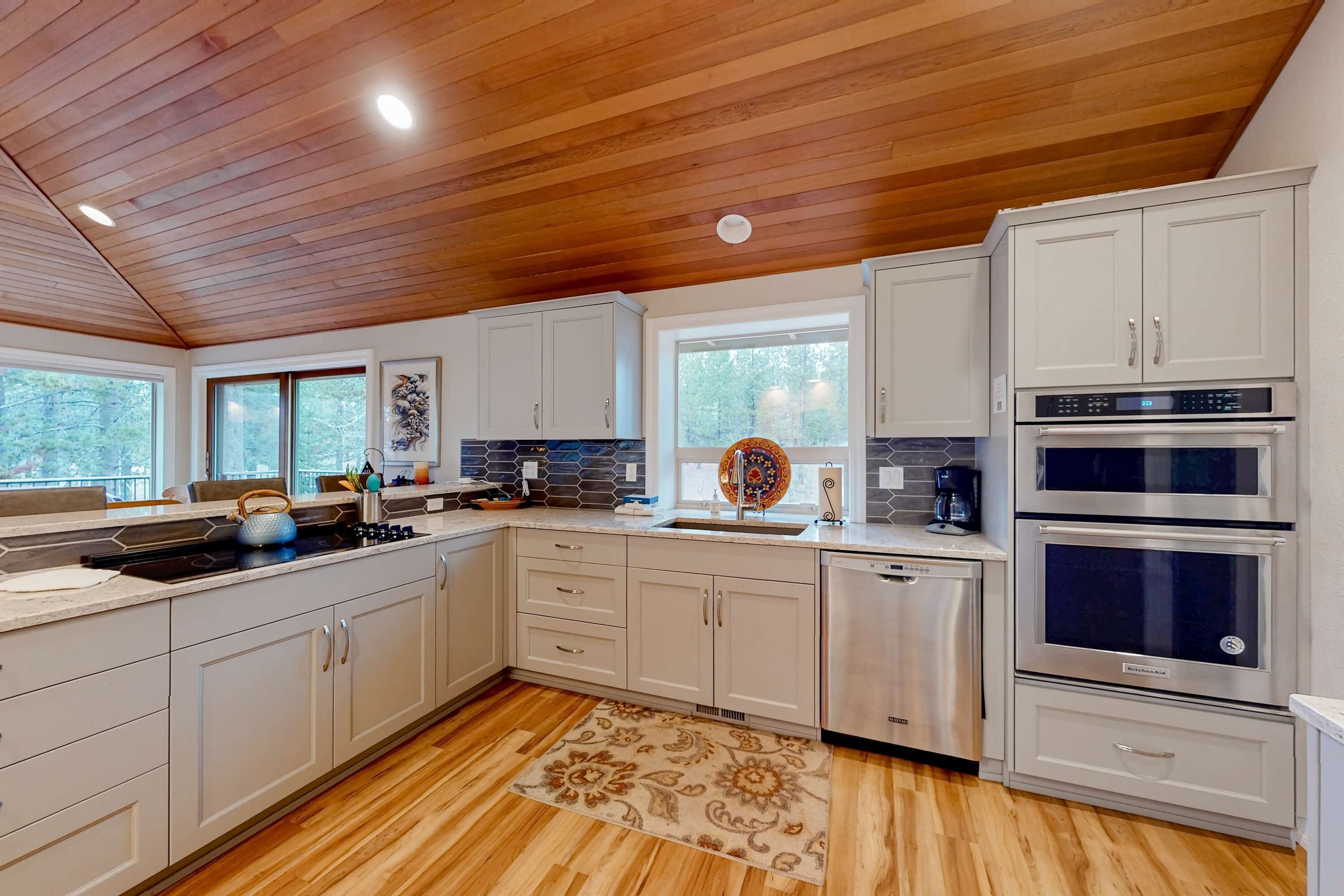 A modern kitchen with wooden ceiling, white cabinets, stainless steel appliances, and a large window.