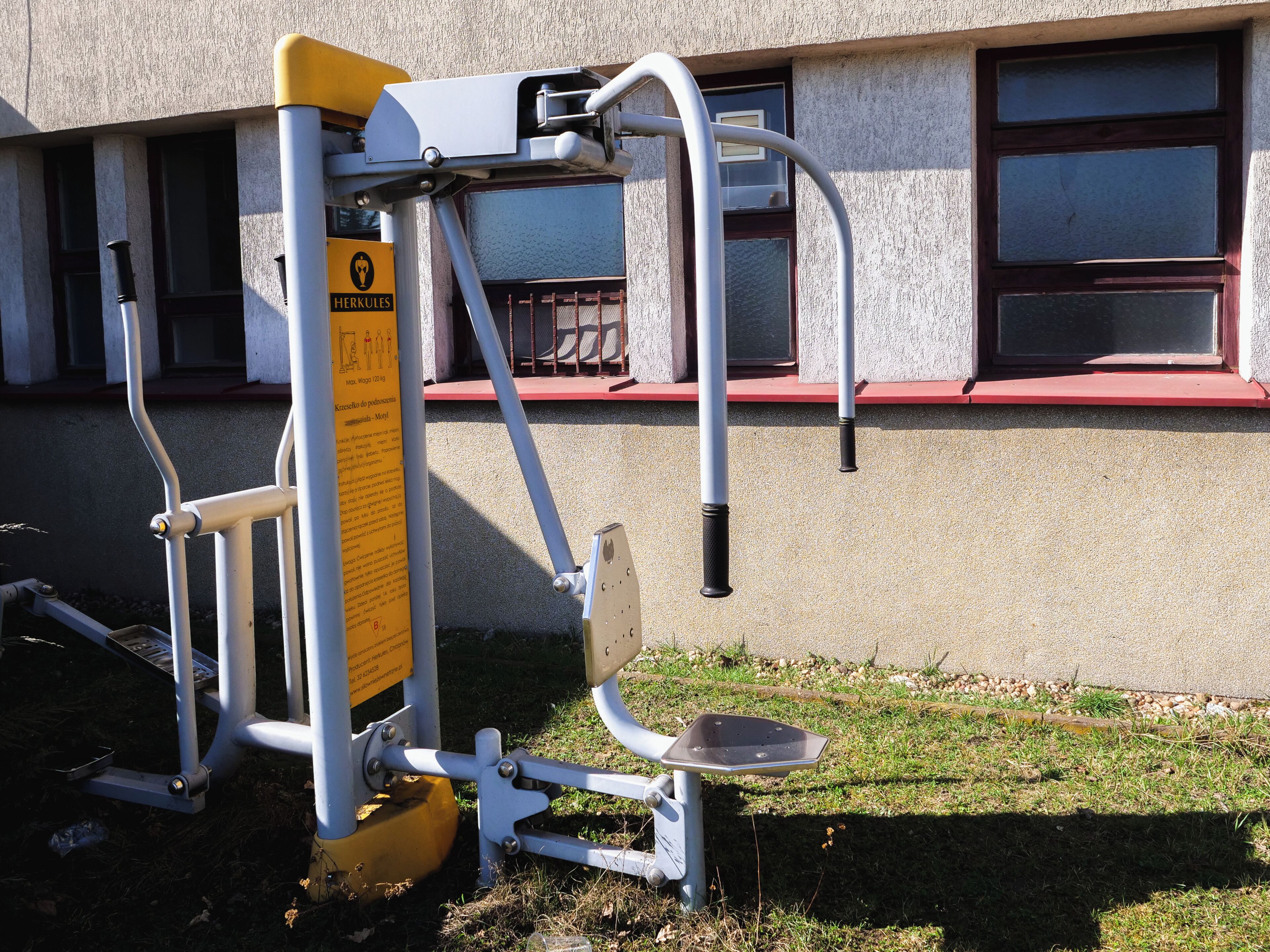 An outdoor exercise machine with a safety sign, a building with windows, and a red window sill.