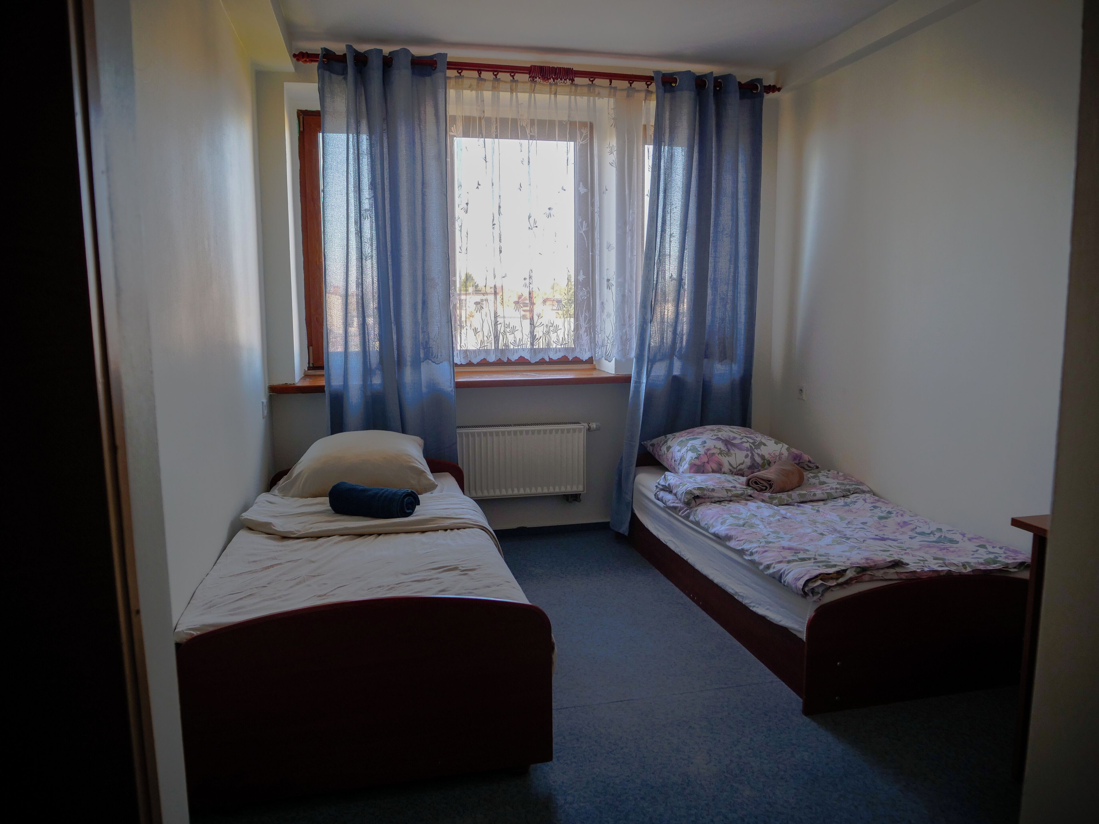 Una habitación pequeña con dos camas individuales, una ventana con cortinas, un radiador y un cabecero de madera.