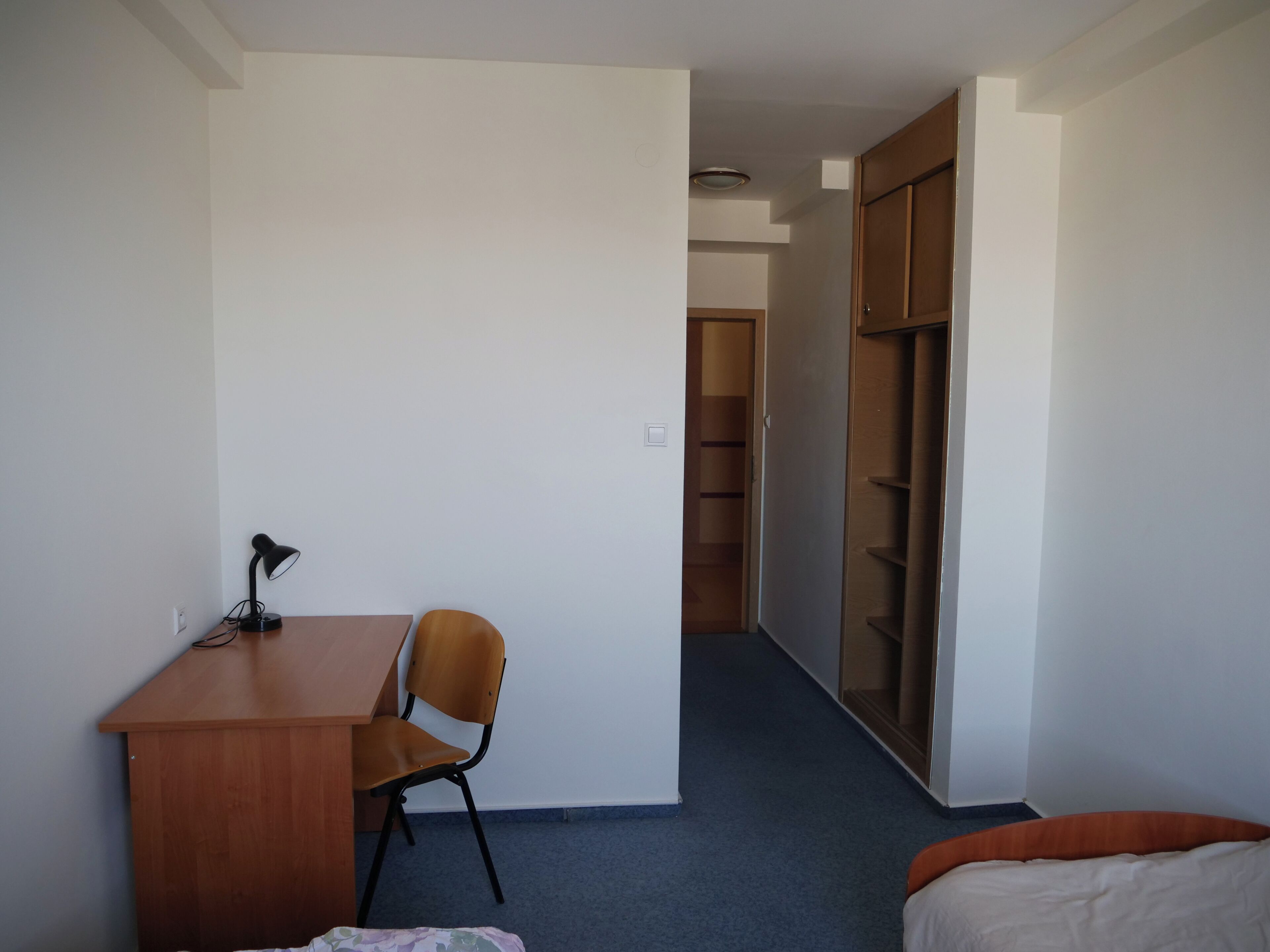 A room with a desk, chair, and a bed. There is a wardrobe and a door leading to another room.