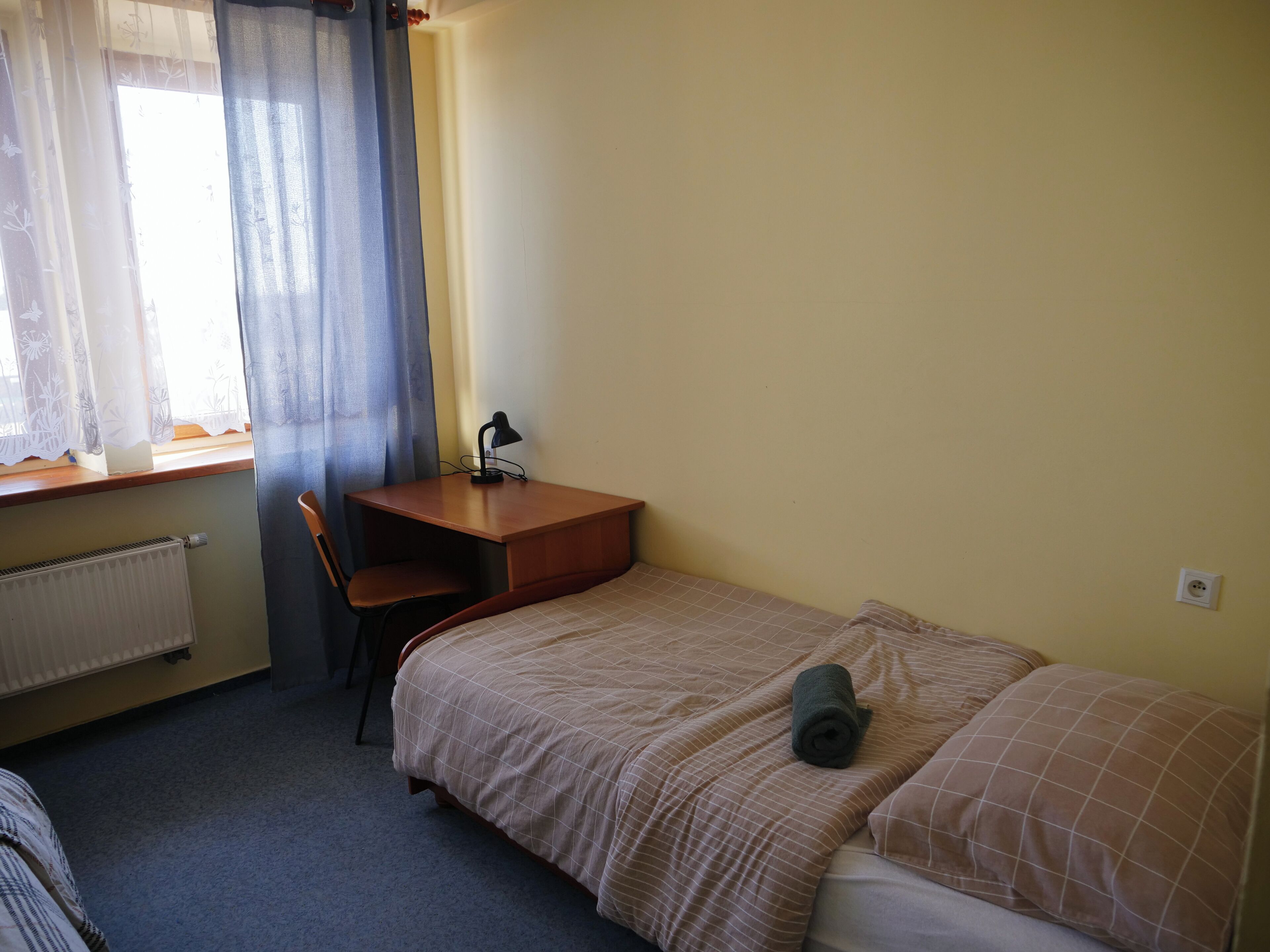 A small, simple room with a bed, a desk, and a chair. There is a radiator on the wall and a window with curtains.
