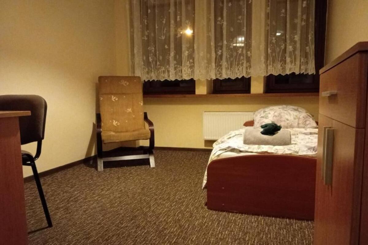 Une chambre d’hôtel avec un lit simple, une chaise, un radiateur et une fenêtre avec un rideau.