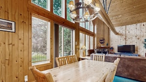 A wooden interior with a dining table, chairs, a chandelier, a TV, and a view through large windows. - Log decor home with private hot tub, deck & shared pools & tennis (Eagle Crest)