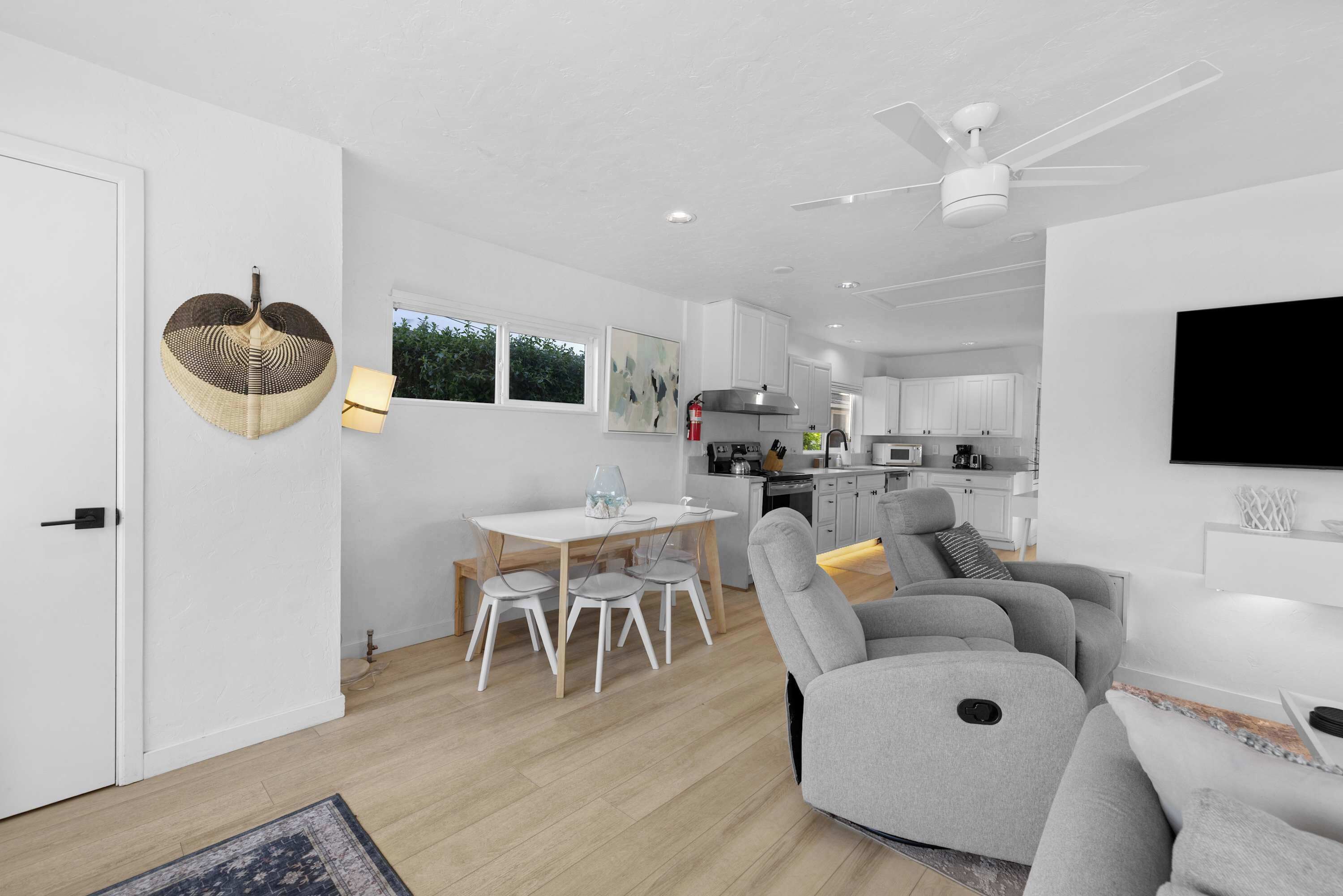 A modern living room with a grey sofa, a dining area with a white table and chairs, a kitchen with white cabinets, and a ceiling fan.
