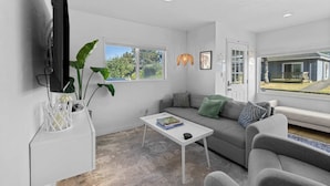 A modern living room with a sofa, a coffee table, a TV, and a window with a view of a house. - Cozy Seaside Bungalow, Brand New Hot Tub, Fast WiFi & W&D - Walk to the Beach (Seaside)