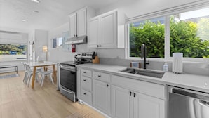 A modern kitchen with white cabinets, stainless steel appliances, a dining area with a table and chairs, and a window with a view of greenery. - Cozy Seaside Bungalow, Brand New Hot Tub, Fast WiFi & W&D - Walk to the Beach (Seaside)
