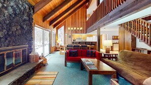A spacious living area with a stone fireplace, wooden ceiling beams, a red sofa, and a staircase. - Beautiful and spacious family-friendly home with indoor hot tub, fireplace, patio (Black Butte Ranch)