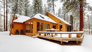 A wooden cabin with large windows, a deck, and a snow-covered roof, surrounded by tall trees. - Beautiful and spacious family-friendly home with indoor hot tub, fireplace, patio (Black Butte Ranch)
