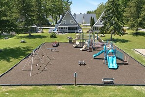 Ein Spielplatz mit Rutschen und Klettergerüsten, umgeben von Gras und Bäumen, mit Häusern im Hintergrund.