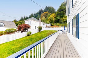 Une terrasse en bois, une clôture blanche en piquets, une pelouse et un bâtiment blanc avec des volets bleus.