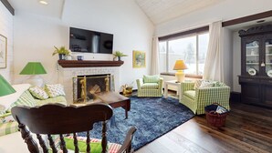 Una sala de estar con chimenea, un televisor montado encima, un sofá verde a cuadros, una mesa de centro de madera y una alfombra azul.