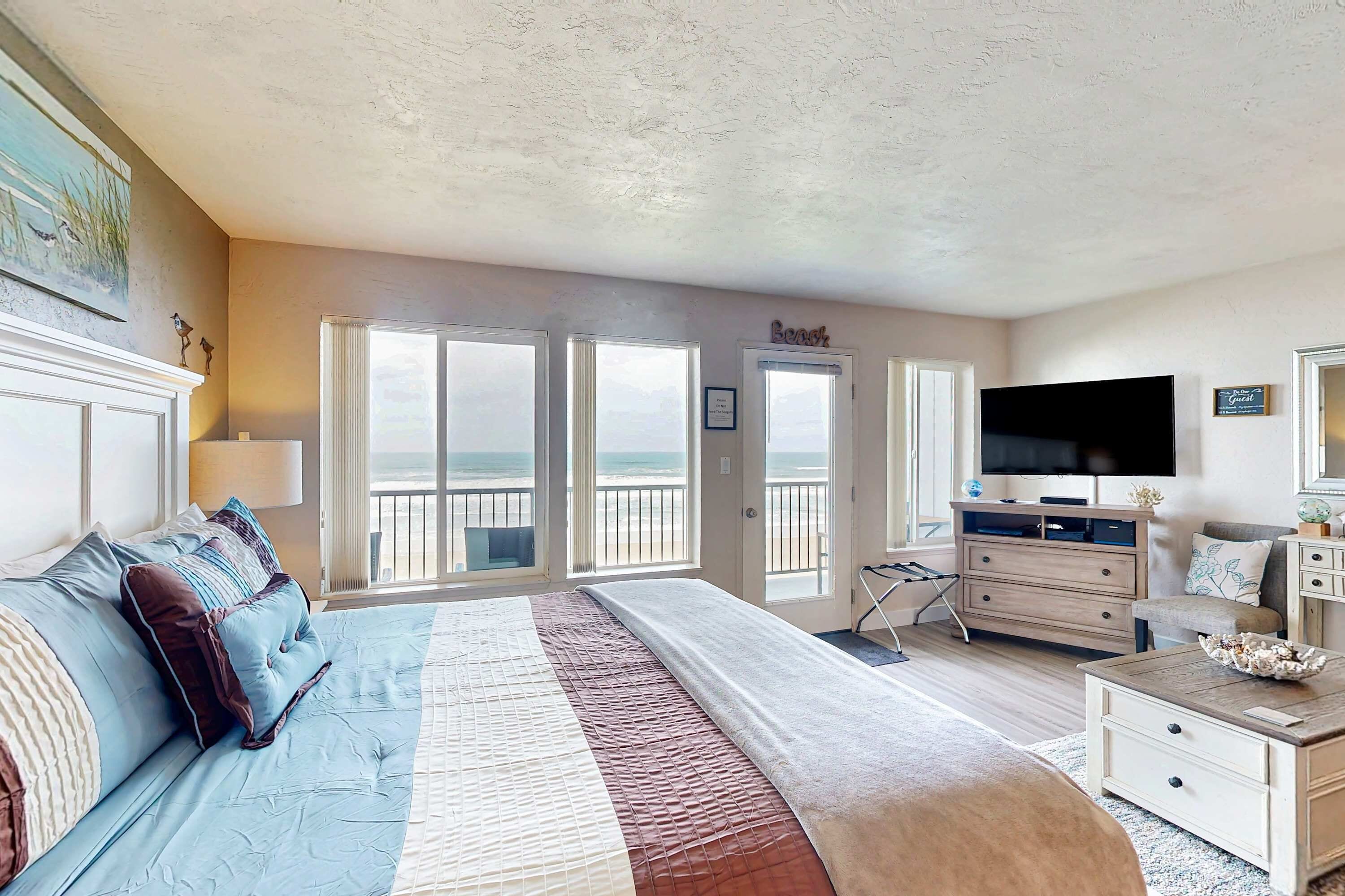 Une chambre avec un grand lit, une commode, une télévision et une vue sur la plage depuis la porte.