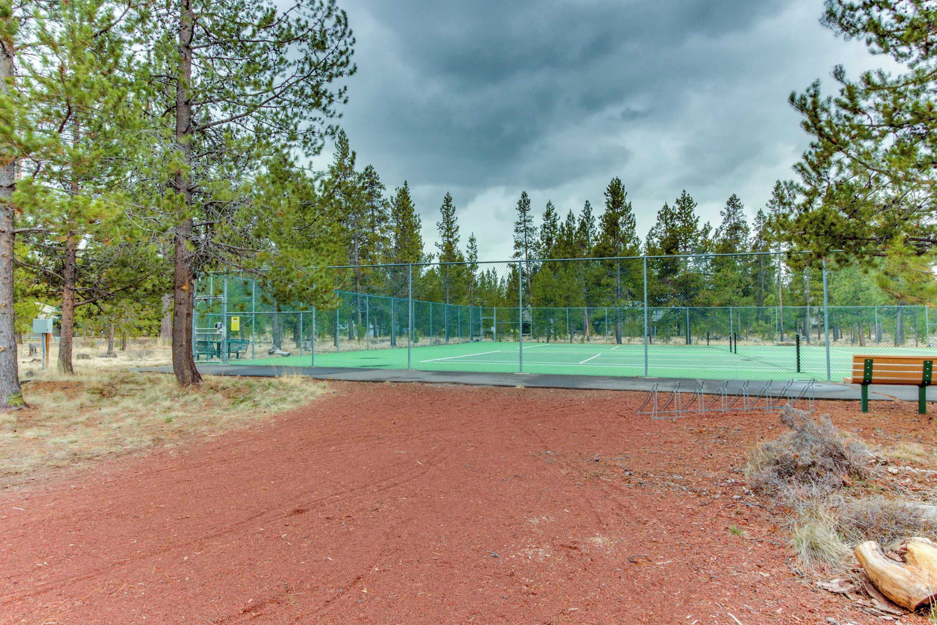 A tennis court with a green fence, a bench, and trees in the background.