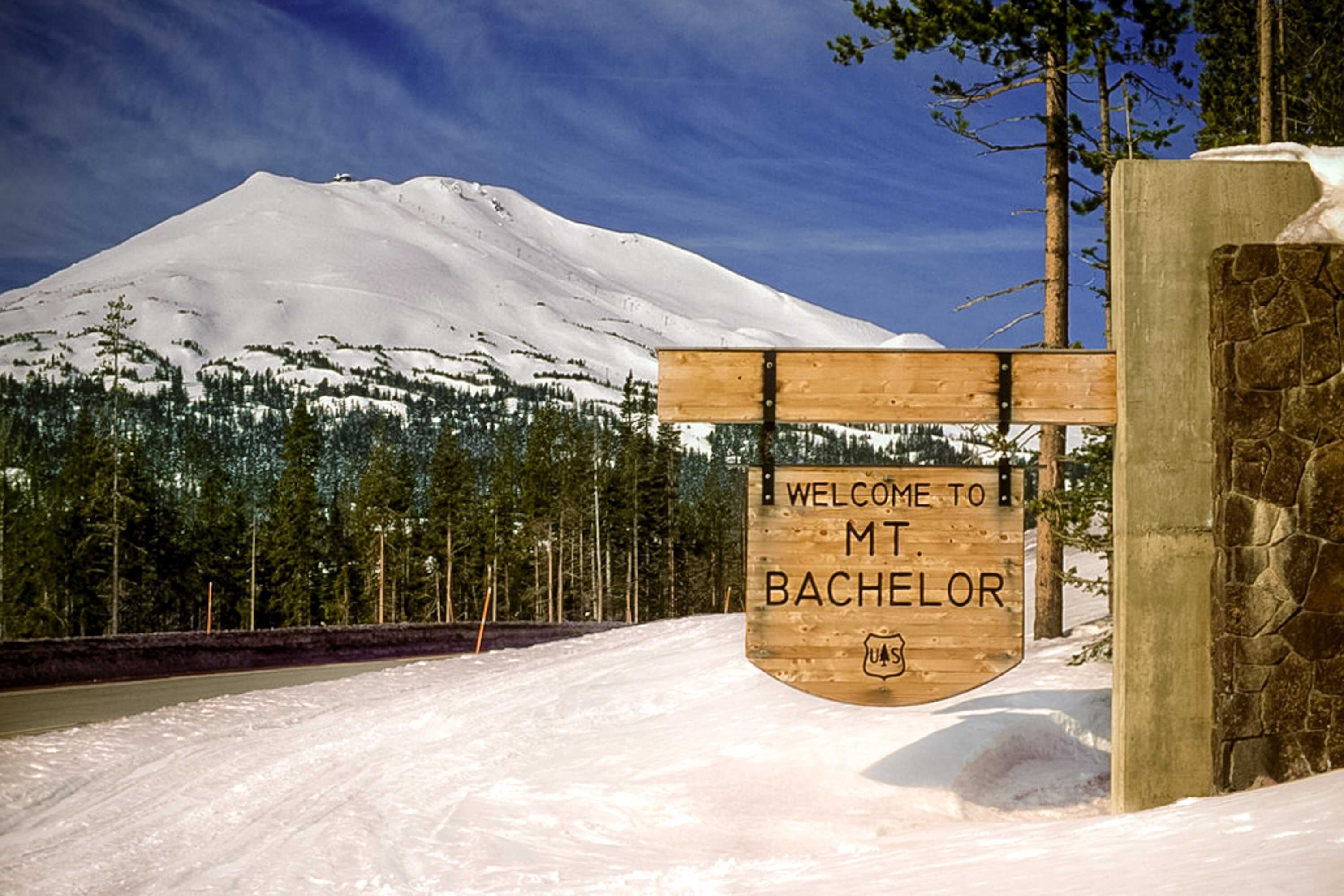 Un cartello di legno con la scritta 'WELCOME TO MT. BACHELOR' e un logo, sullo sfondo di una montagna innevata.