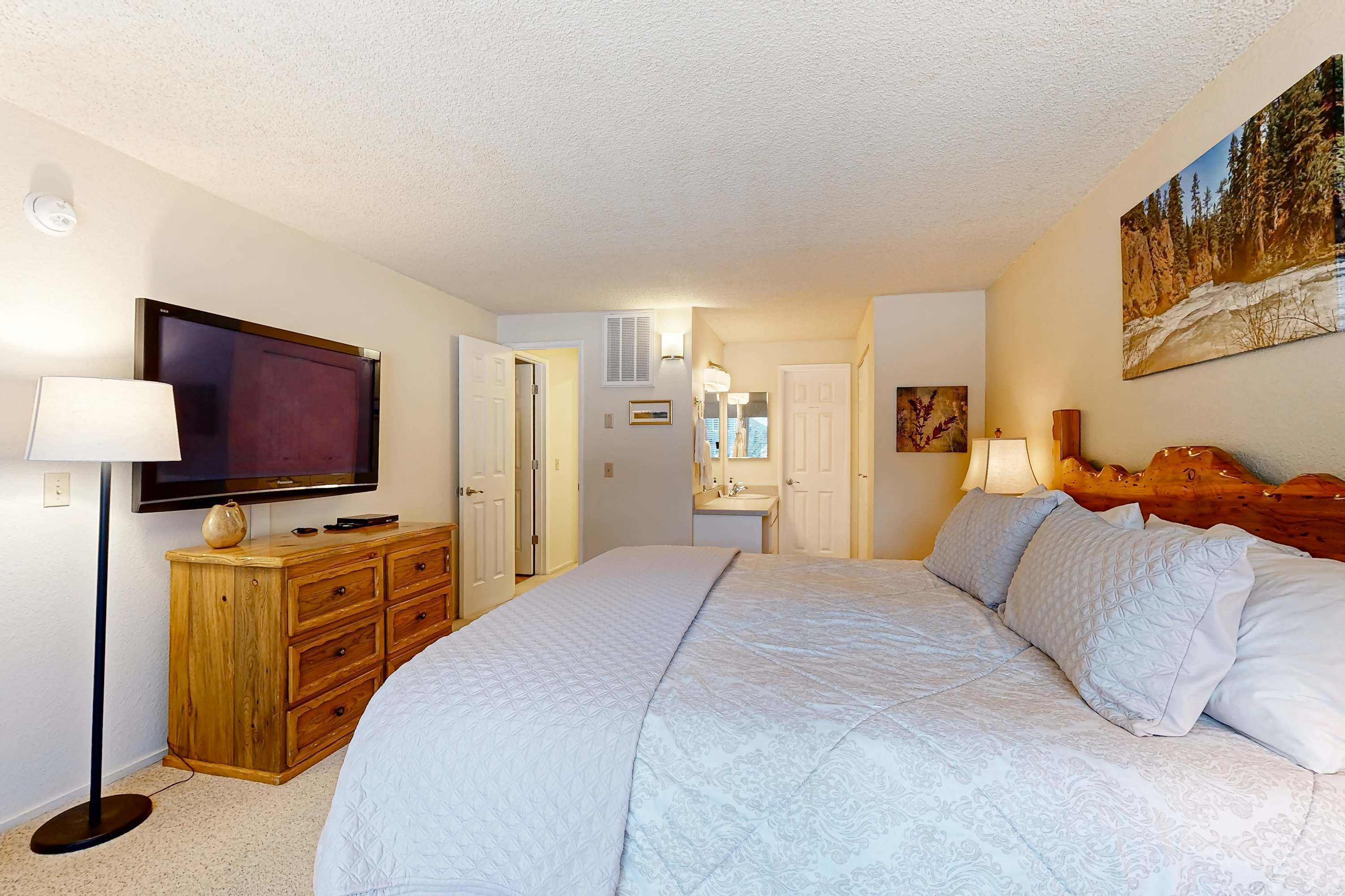 Une chambre avec un grand lit, un téléviseur posé sur une commode en bois, une salle de bain avec un lavabo et un miroir, et un tableau accroché au mur.