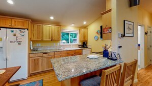 A kitchen with wooden cabinets, granite countertops, a built-in island with seating, a refrigerator, and a window with a view of greenery. - Adorable dog-friendly home with gas grill, gas fireplace & private back patio (Manzanita)
