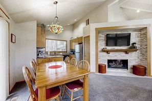 A dining area with a wooden table and chairs, a kitchen in the background, a stone fireplace, and a hanging light fixture.