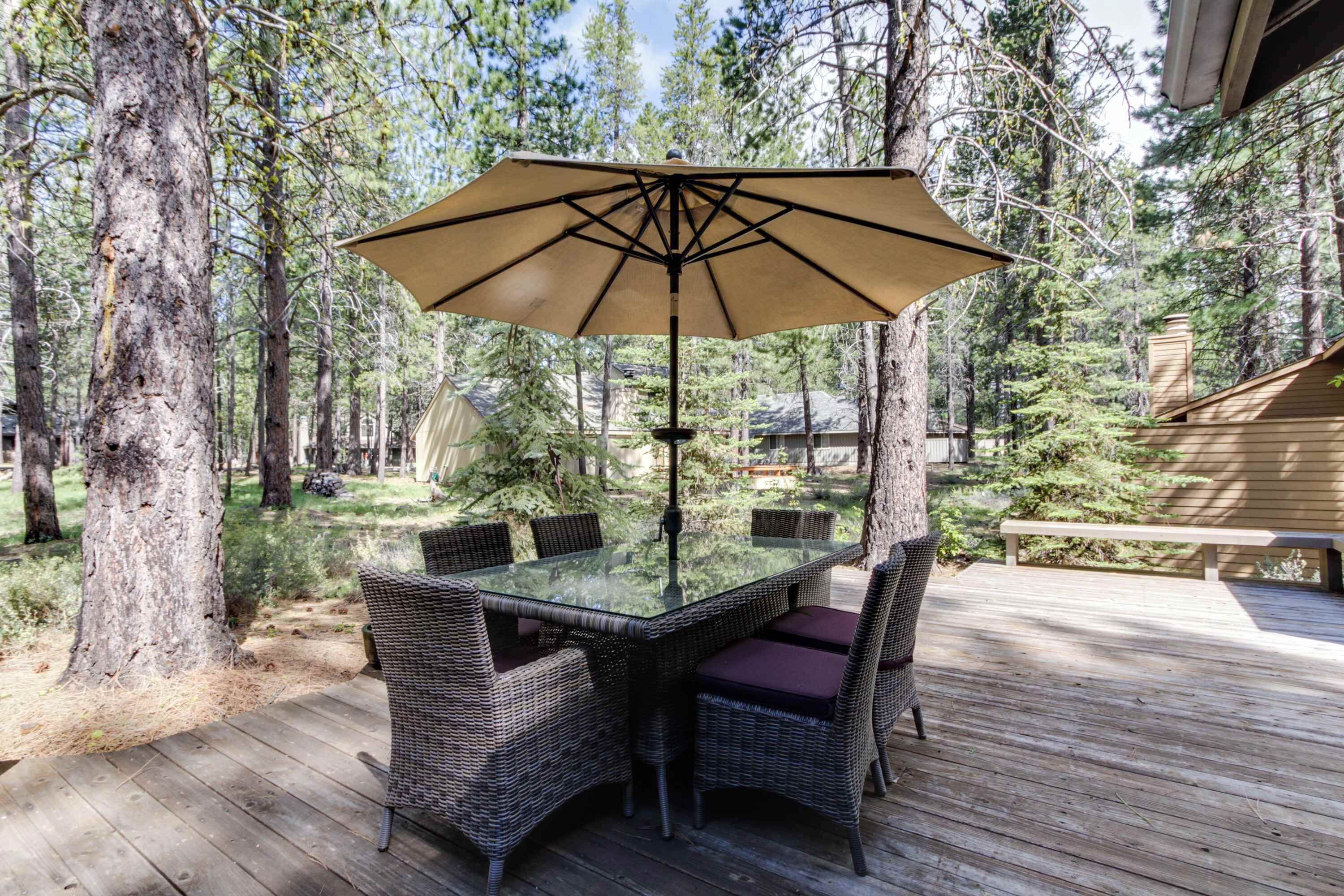 A glass-top table with wicker chairs and a beige umbrella in a forested outdoor area.