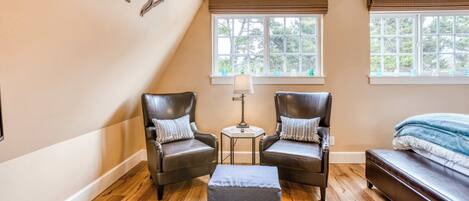 A room with two leather chairs, a small table with a lamp, a bed, and a window with blinds.