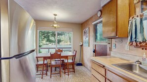 Una cocina con refrigerador, mesa de madera, sillas, una ventana con persianas, un escoba y un lavamanos.