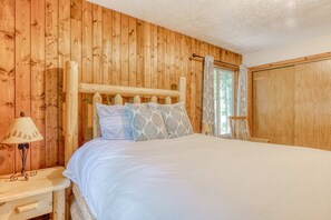 A wooden-paneled room with a bed, a nightstand, and a window with curtains. - Serene Cottage on the River with Firepit, Deck, and Bunk Room (Rhododendron)