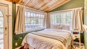 Un dormitorio con techo de madera, paredes verdes, una cama con colcha estampada, dos ventanas con cortinas y una mesita de noche con un libro.