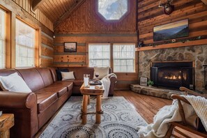A log cabin interior with a stone fireplace, leather sofa, and a large window. - Beautiful Cabin, Perfect Location. Book Now! Minutes From Town! (Sevierville)
