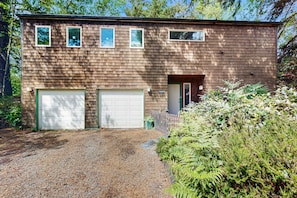 A two-story building with a garage, a small porch, and a gravel driveway. - Forest-view home with fireplace, deck - Walk to the Beach & Golf Course (Manzanita)