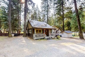 A cabin with a porch, surrounded by tall trees, and a small adjacent building.