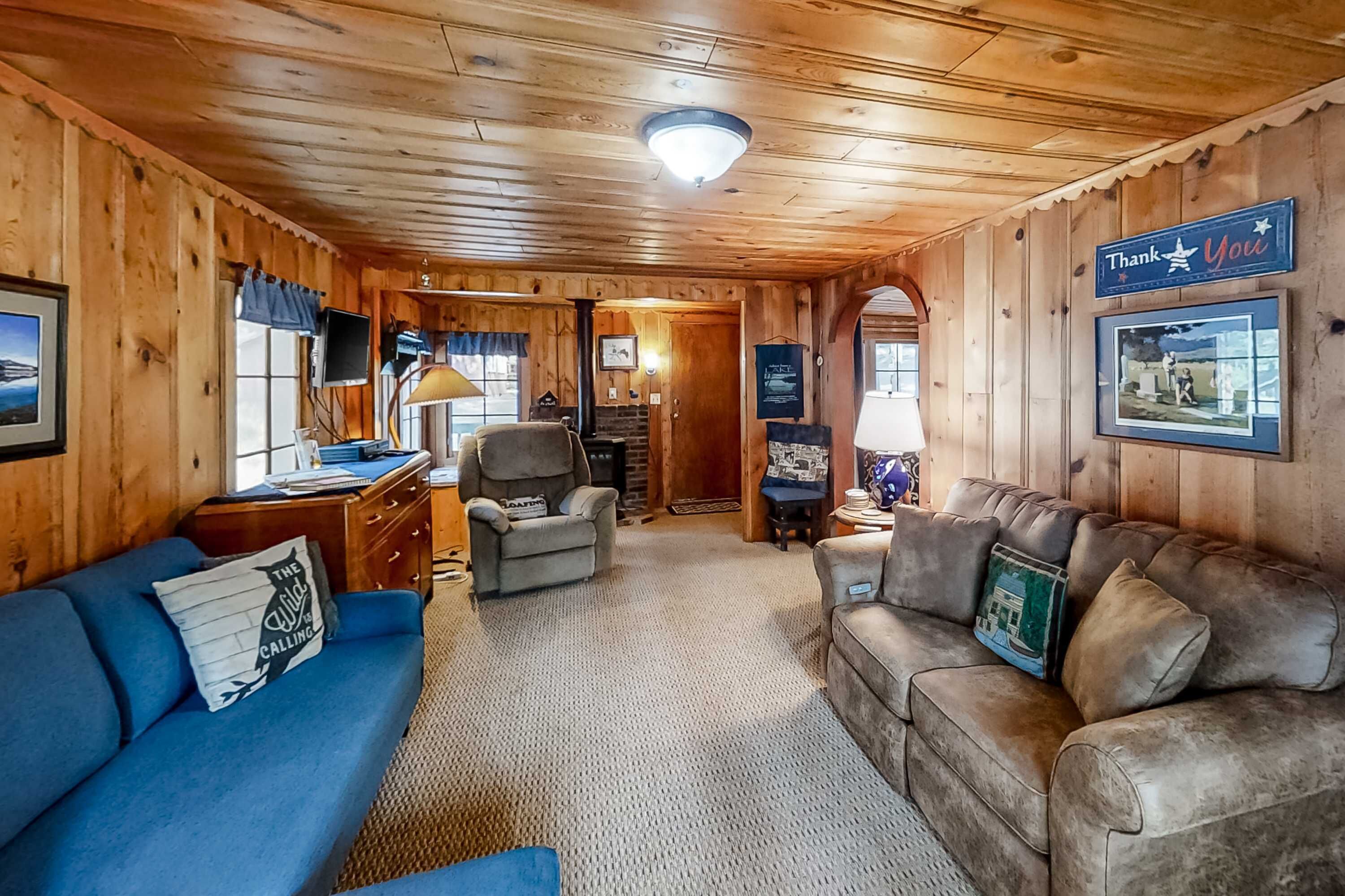 A cozy cabin interior with wooden walls, a blue sofa, a leather couch, a desk with a lamp, and a 'Thank You' sign.