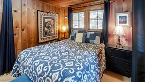 Ein Schlafzimmer mit Holzwand, einem Bett, zwei Nachttischlampen, einem gerahmten Bild an der Wand und einem Fenster mit Vorhängen.