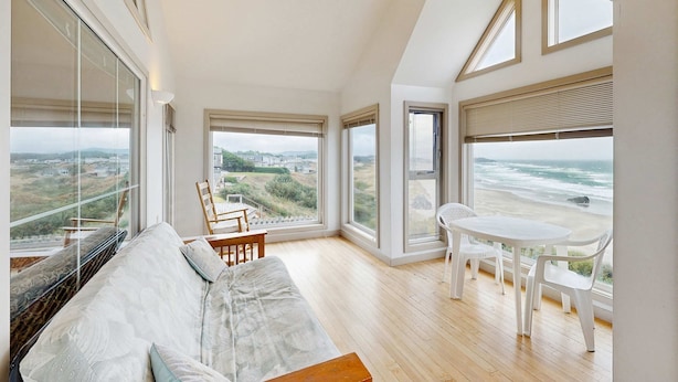 Een ruime kamer met een groot raam met uitzicht op het strand, een witte bank en een kleine eettafel met stoelen.