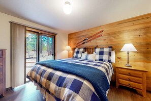 A bedroom with a large bed, wooden headboard, skis mounted on the wall, and a sliding glass door leading outside. - Loft home with private hot tub, deck, grill, fireplace, & 10 SHARC passes (Sunriver)