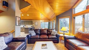 A spacious living area with leather sofas, a coffee table, and large windows offering a view of the outdoors. - Inviting house with hot tub, game tables, bikes, & partial mountain view (Sunriver)