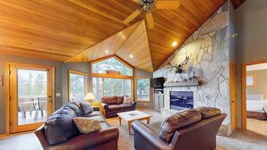 A spacious living room with leather sofas, a stone fireplace, and large windows. - Inviting house with hot tub, game tables, bikes, & partial mountain view (Sunriver)