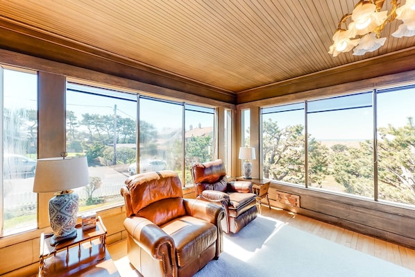 Quarto amplo com grandes janelas, poltronas de couro, uma mesinha com abajur e vista para o exterior.