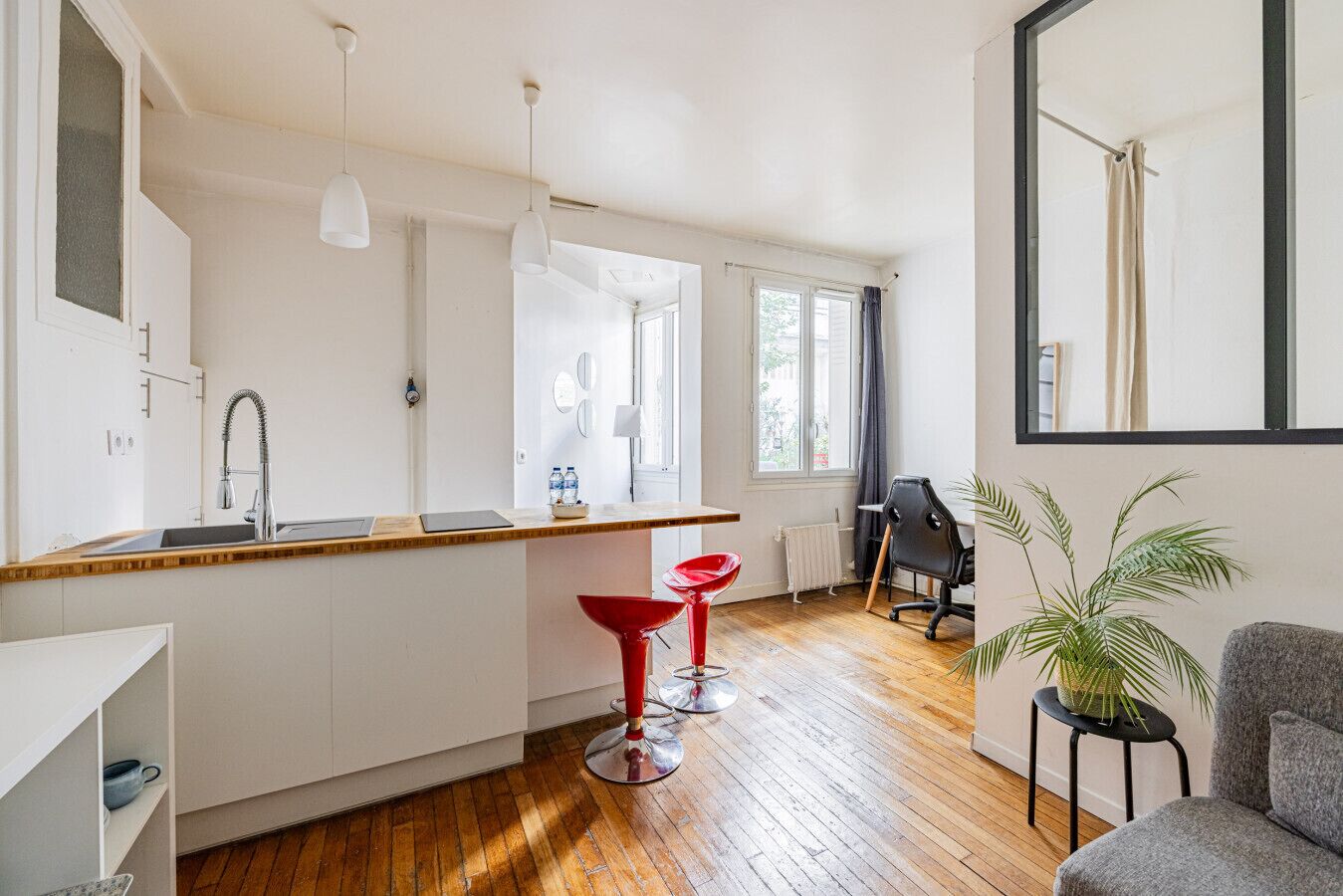 A modern kitchen with a wooden countertop, a sink, and a red chair. There is a black office chair and a grey sofa in the living area.